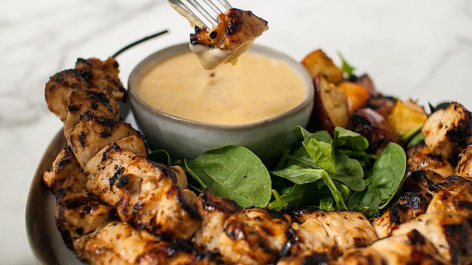 Grilled chicken skewers on a plate with fresh spinach, roasted vegetables, and a bowl of creamy dipping sauce, with a fork holding a piece of chicken above the sauce.