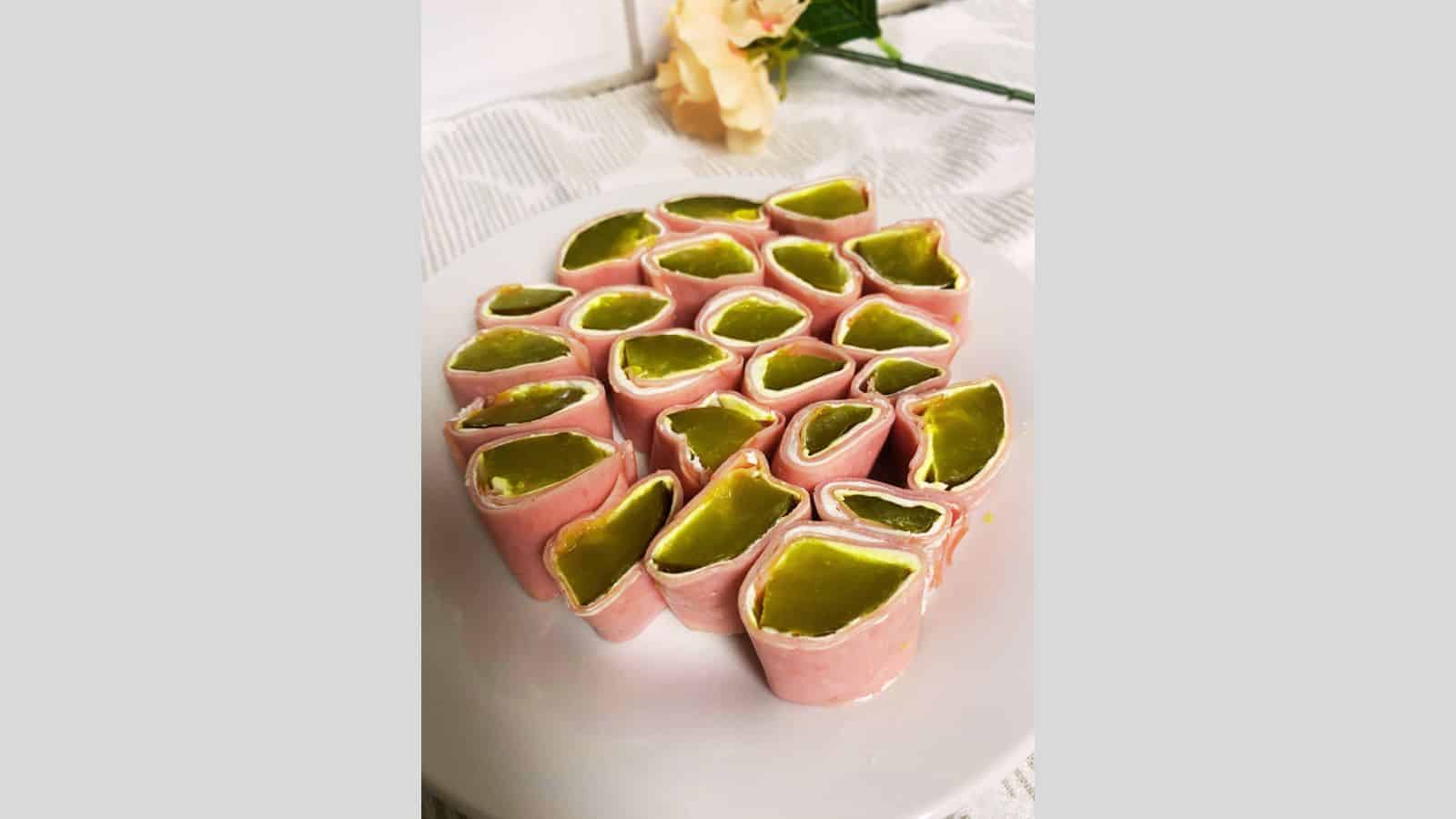 Sliced ham rolls filled with green pickles and cream cheese arranged on a white plate, with a flower in the background.