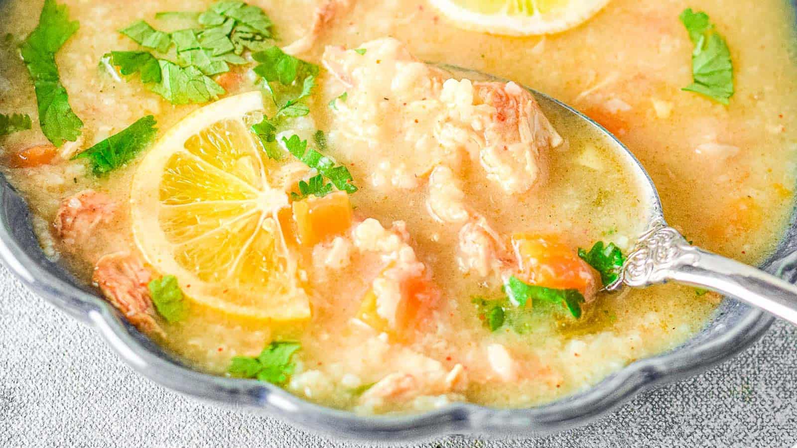 Close-up of a bowl of soup containing rice, shredded chicken, chopped vegetables, lemon slices, and fresh cilantro, with a spoon scooping up a portion.