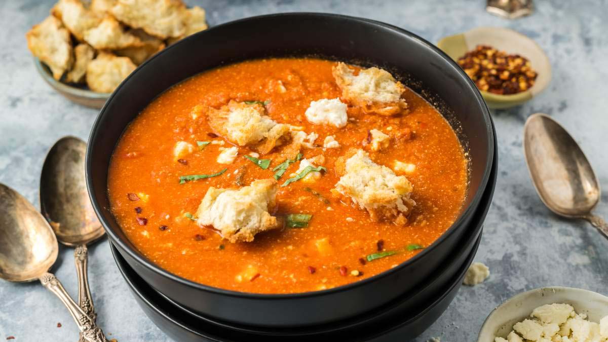 A bowl of tomato soup topped with crumbled bread and herbs, surrounded by spoons, bread pieces, and a small dish of red pepper flakes.