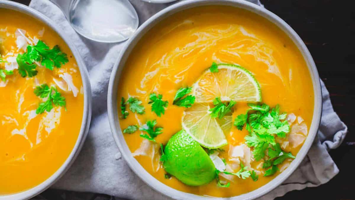 A bowl of creamy orange soup garnished with lime slices, fresh cilantro, and coconut flakes, set on a gray cloth next to another bowl.
