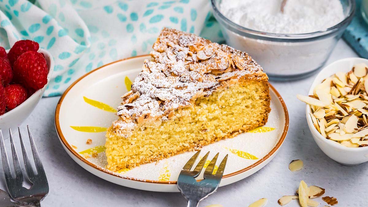 A slice of almond cake topped with powdered sugar and almond flakes on a plate, surrounded by raspberries, sliced almonds, and a bowl of powdered sugar.