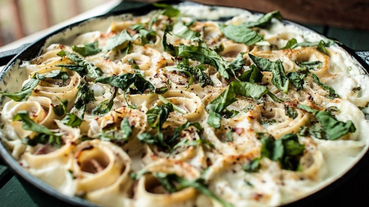 A close-up of a baked pasta dish topped with fresh spinach, creamy sauce, and seasonings, served in a black pan.