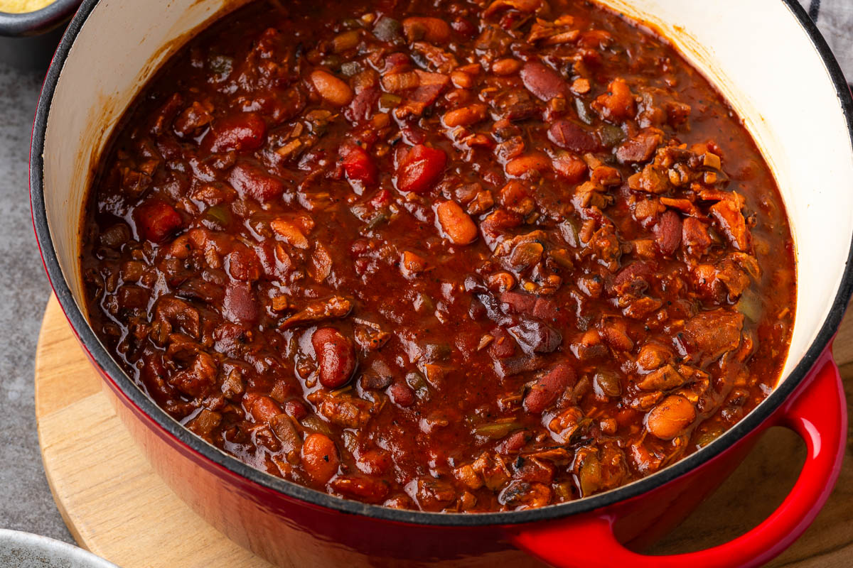 A close-up of a red pot filled with thick, hearty chili containing beans and vegetables in a rich, reddish-brown sauce.