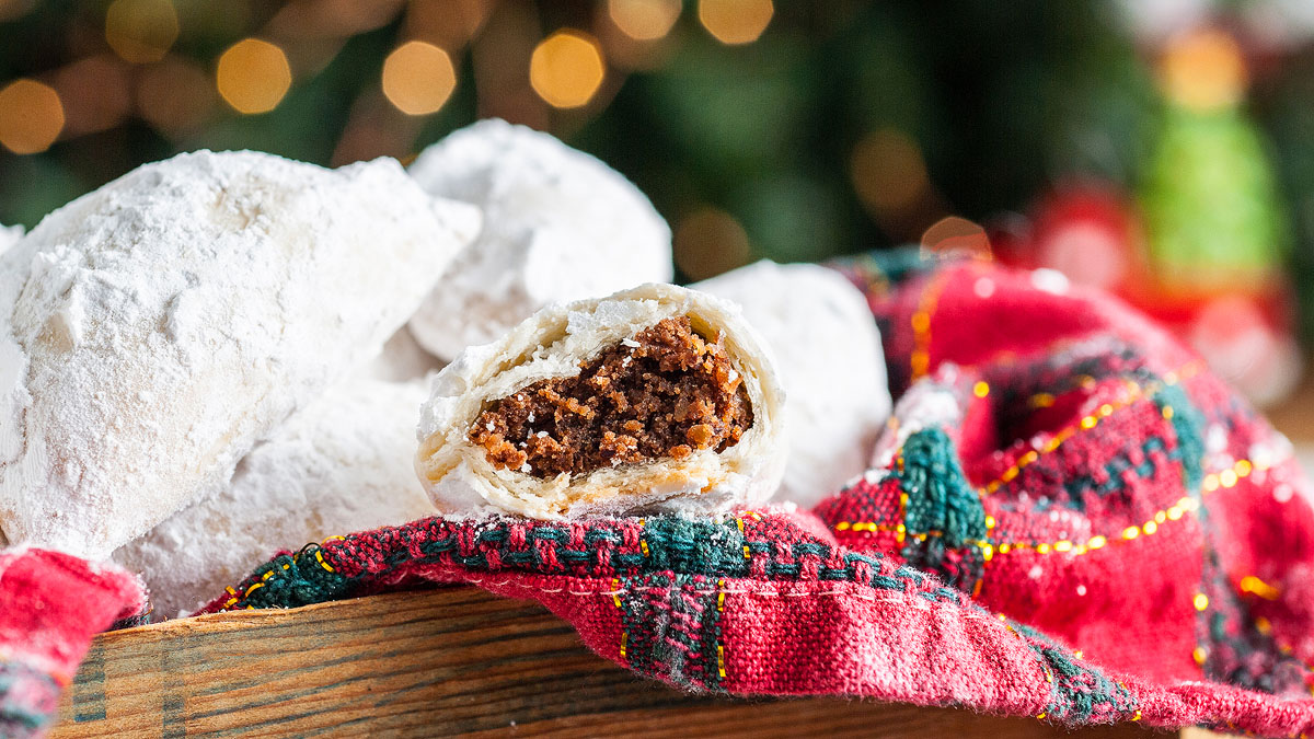 Powdered sugar-coated cookies, one cut open to show a brown filling, rest on a red and green cloth with blurred holiday lights in the background.