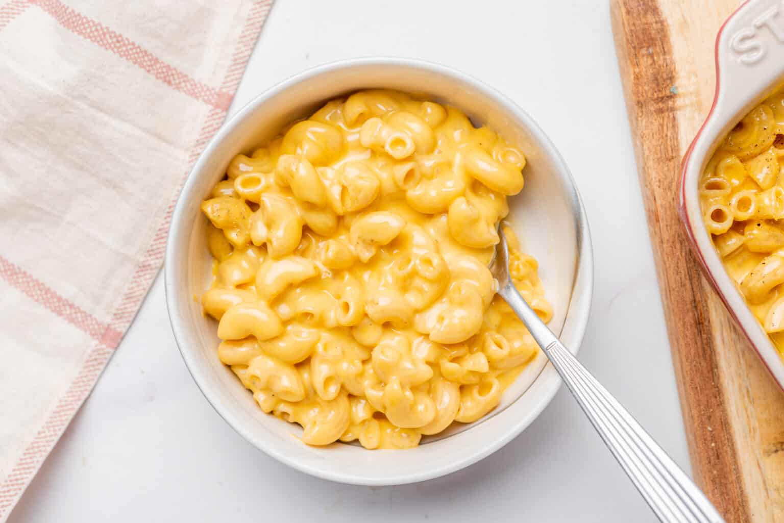A bowl of macaroni and cheese with a fork, placed on a white surface next to a striped cloth and a wooden board.