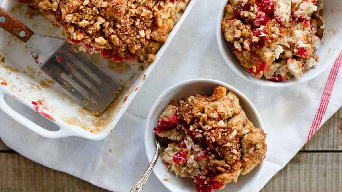 A baking dish and two bowls of fruit crumble with oat topping, one bowl has a spoon, set on a wooden surface with a red-striped napkin.