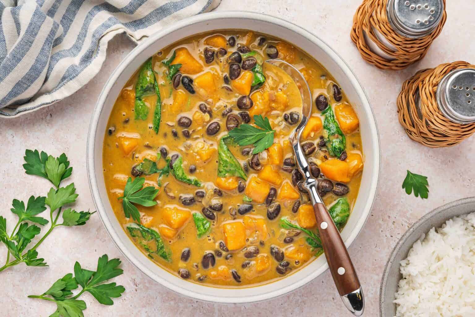A bowl of curry with black beans, sweet potatoes, spinach, and a creamy sauce, garnished with parsley. A serving spoon rests in the bowl; rice and seasonings are nearby.