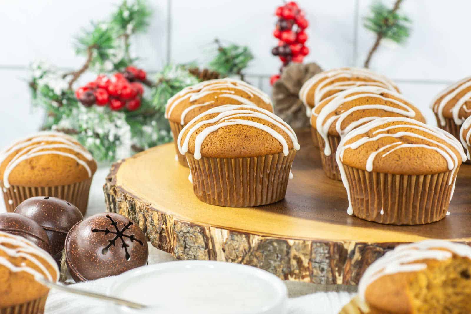 Gingerbread muffins with white icing are arranged on a wooden tray, with Christmas decorations and a bowl of icing in the background.