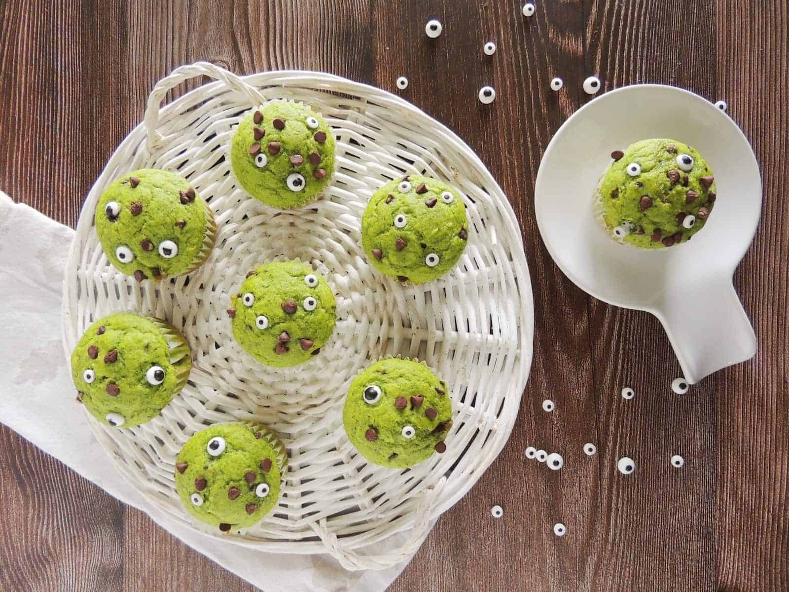 Green muffins decorated with candy eyes and chocolate chips are arranged in a white basket on a wooden surface, with one muffin placed in a white dish nearby.