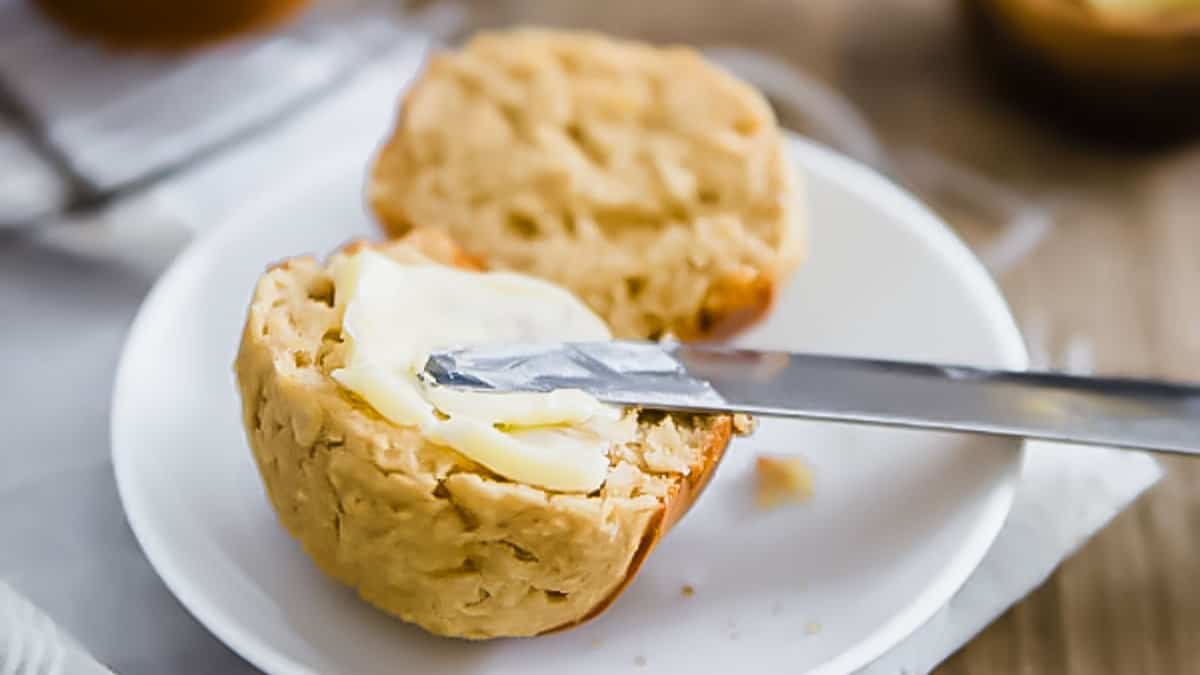 A biscuit split in half on a white plate, with a knife spreading butter on one half.