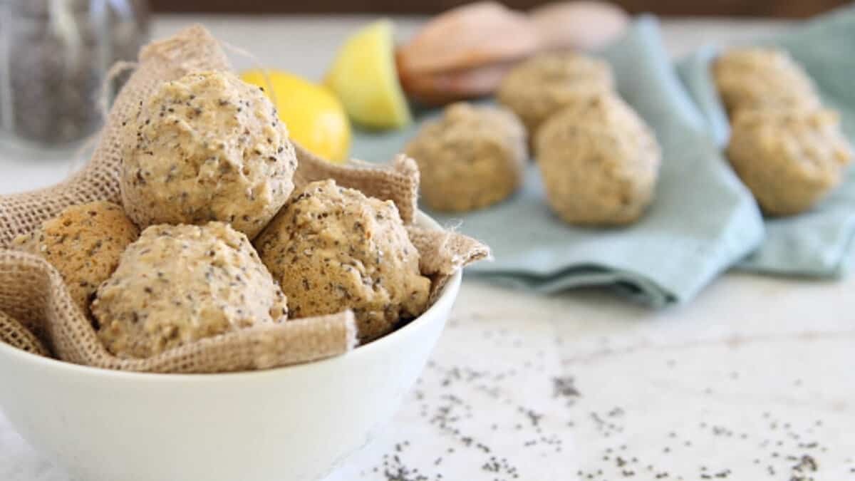 A white bowl lined with burlap contains round, chia seed-studded baked rolls; more rolls are on a blue cloth in the background.