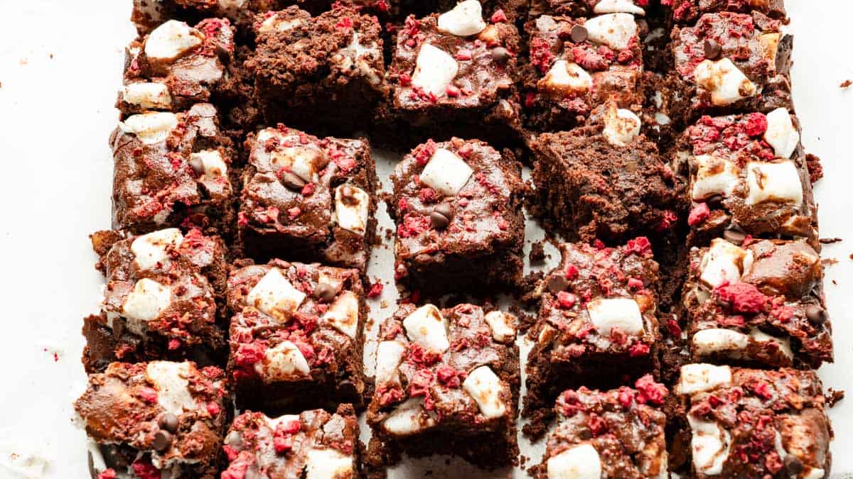 Rows of chocolate brownies topped with marshmallows and pieces of red berry, cut into squares and arranged on a white surface.