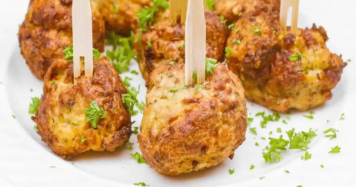 Several browned meatballs garnished with chopped parsley are arranged on a white plate, each pierced with a small wooden pick.