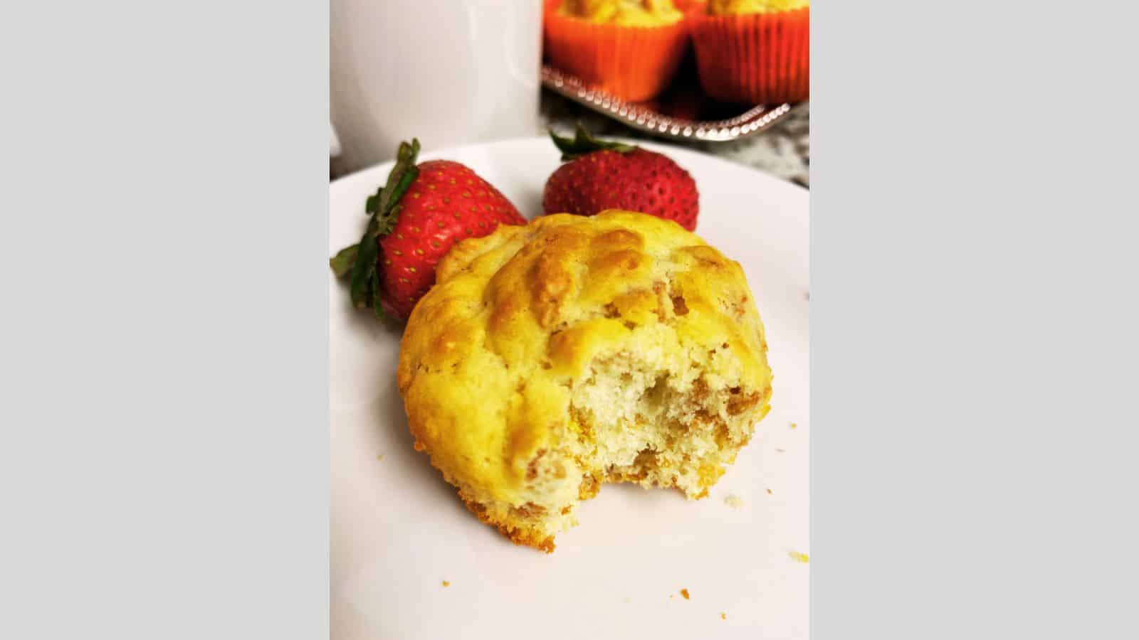 A partially eaten muffin on a white plate with two strawberries, a mug, and more muffins in the background.