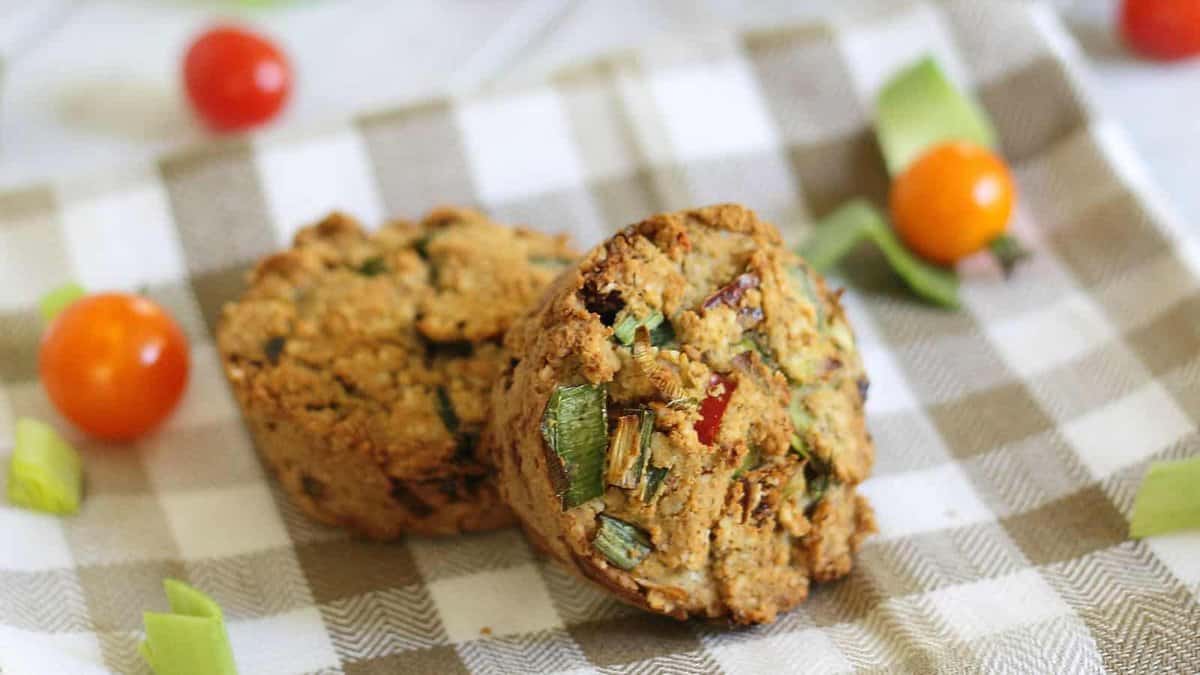 Two savory muffins with visible chopped vegetables rest on a checkered cloth, surrounded by cherry tomatoes and green vegetable pieces.