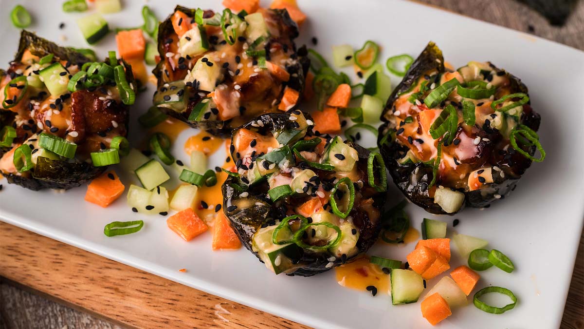A plate of bite-sized sushi topped with diced vegetables, green onions, sesame seeds, and drizzled sauce, served on a white rectangular dish.