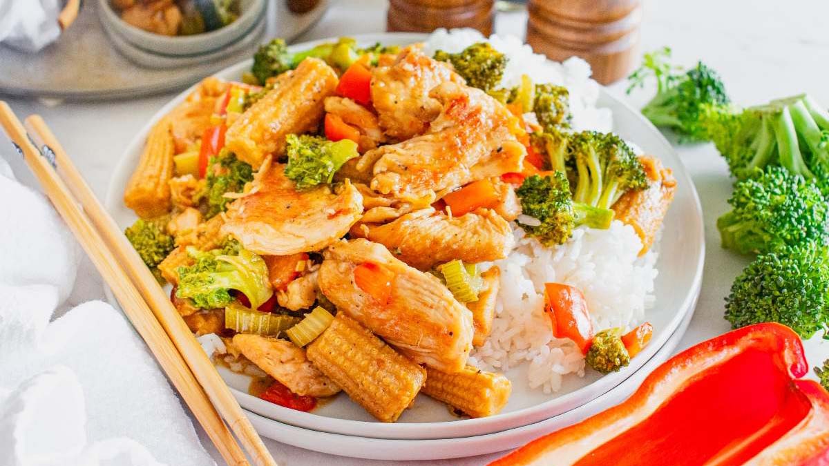A plate of white rice topped with stir-fried chicken, baby corn, broccoli, and red bell peppers, with chopsticks placed on the side.