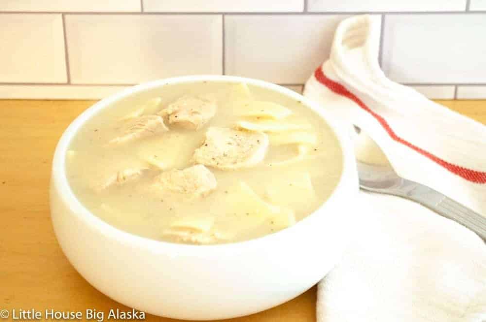 A white bowl of creamy chicken and noodle soup on a wooden surface, with a white towel and spoon beside it.