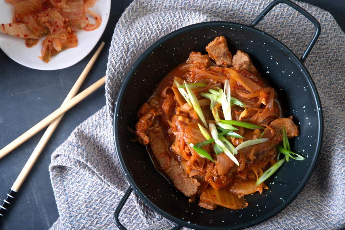 A black bowl with stir-fried pork, onions, and kimchi, garnished with sliced green onions. Chopsticks and a small plate of kimchi are nearby.