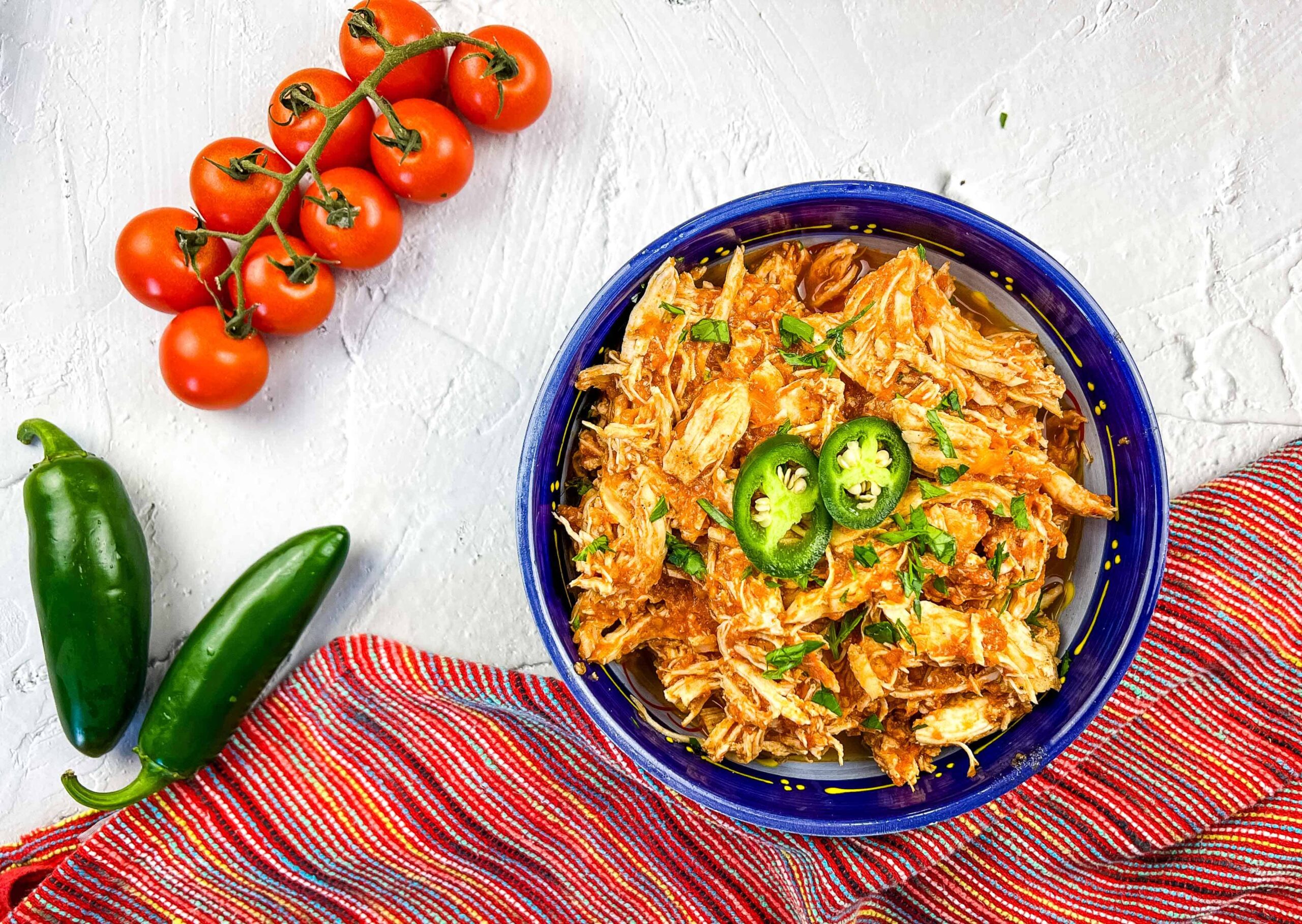 A bowl of shredded chicken with sliced jalapenos and herbs, next to a cluster of cherry tomatoes, jalapenos, and a striped cloth on a white surface.