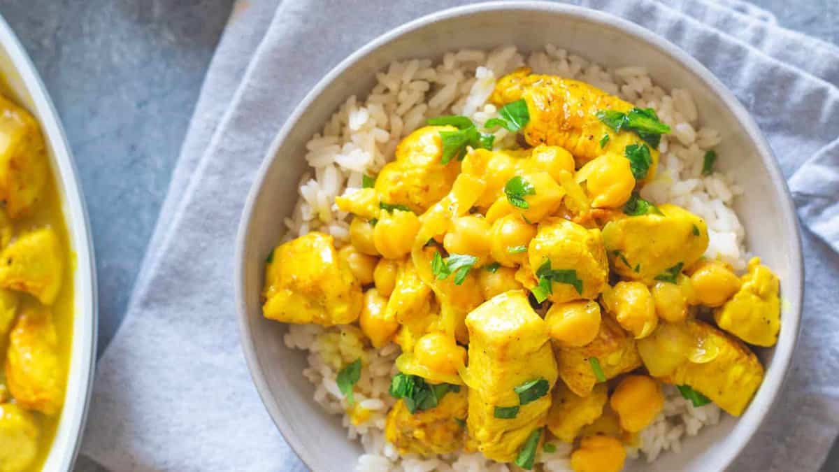A bowl of white rice topped with yellow curry chicken, chickpeas, and chopped herbs, placed on a gray napkin.