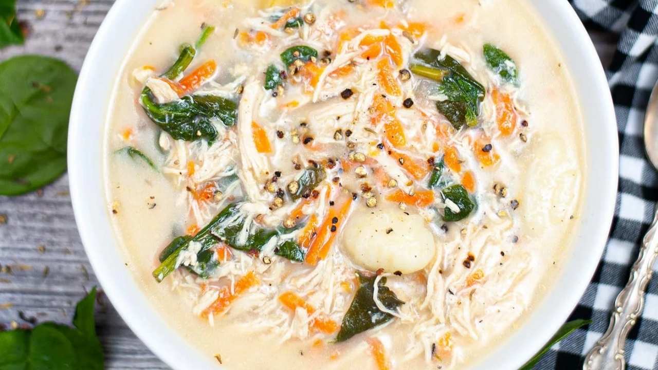 A bowl of creamy chicken and vegetable soup with shredded chicken, carrots, spinach, and gnocchi, garnished with cracked black pepper.