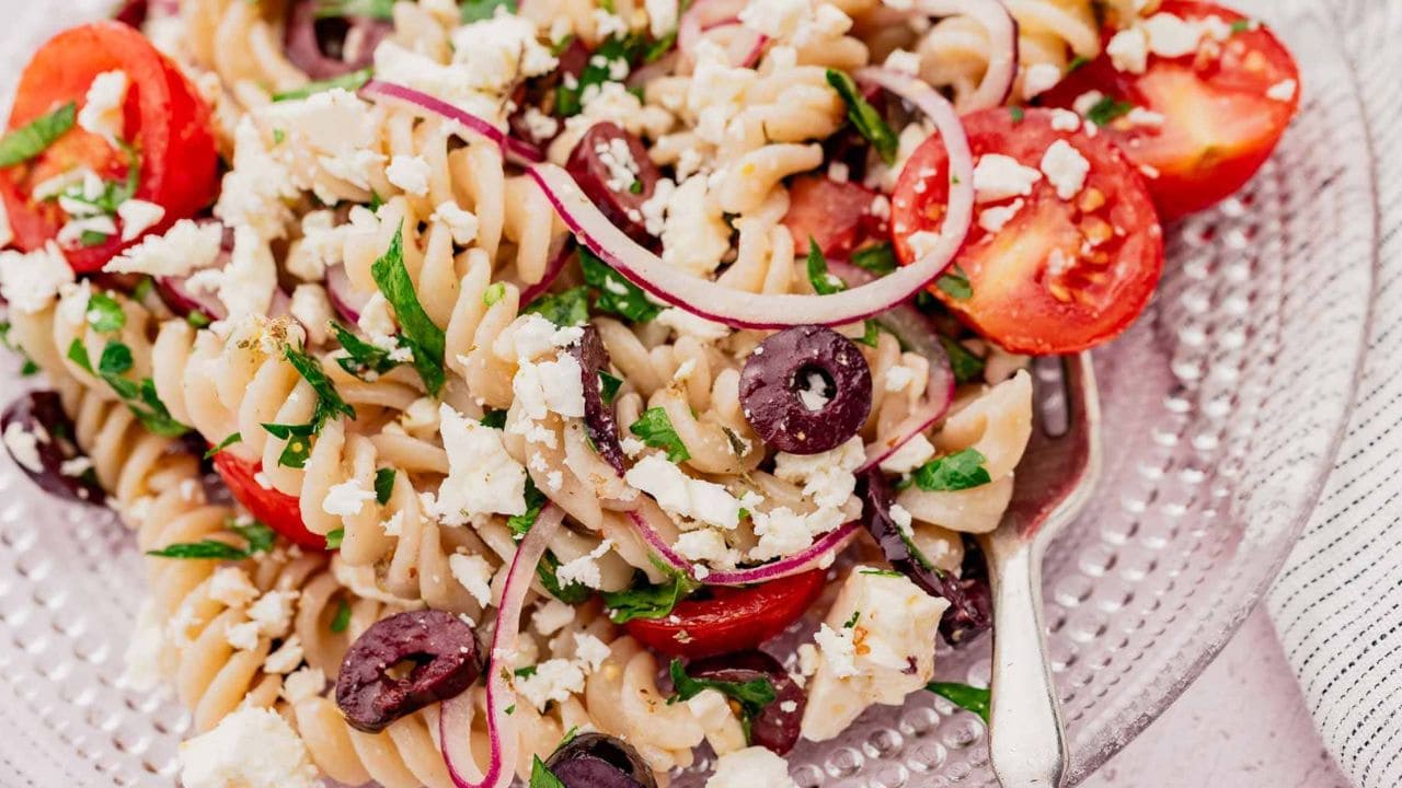 A plate of rotini pasta salad with cherry tomatoes, Kalamata olives, red onion slices, feta cheese, and chopped parsley.