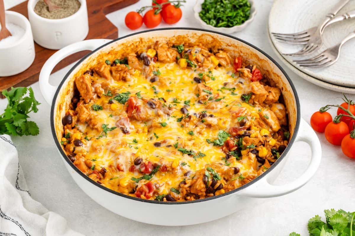 A white cast iron pot filled with a baked dish of rice, black beans, corn, tomatoes, chicken, and melted cheese, garnished with chopped herbs.