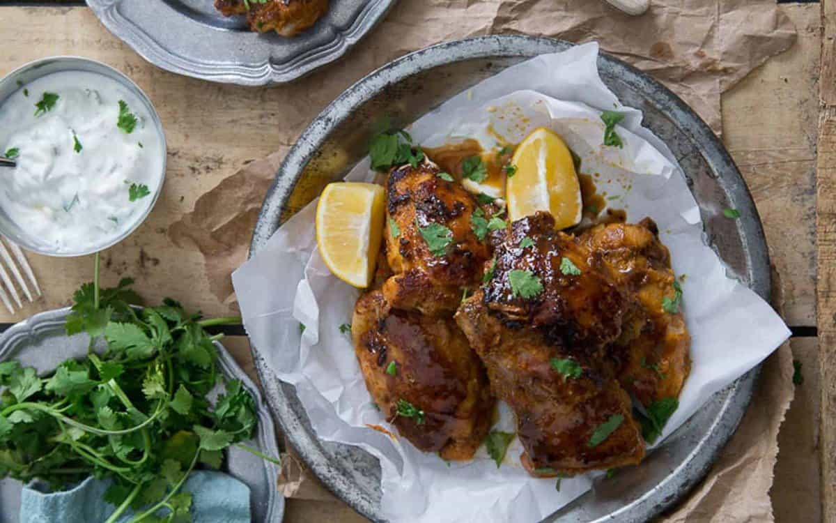Grilled chicken pieces garnished with cilantro and lemon wedges on a metal plate, with a bowl of yogurt sauce and fresh herbs nearby.