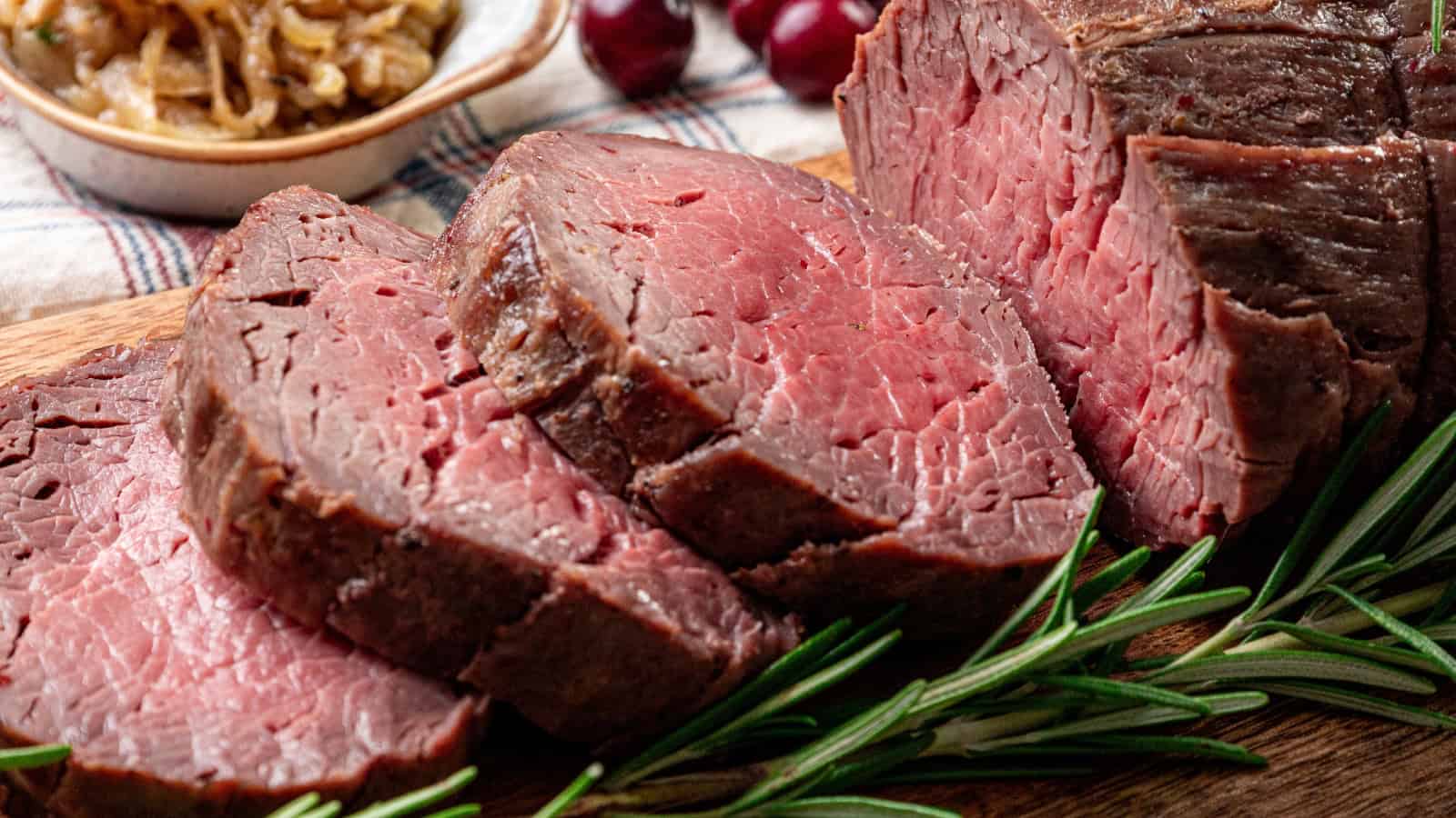 Sliced medium-rare roast beef on a wooden cutting board, garnished with rosemary, with caramelized onions and cherries in the background.