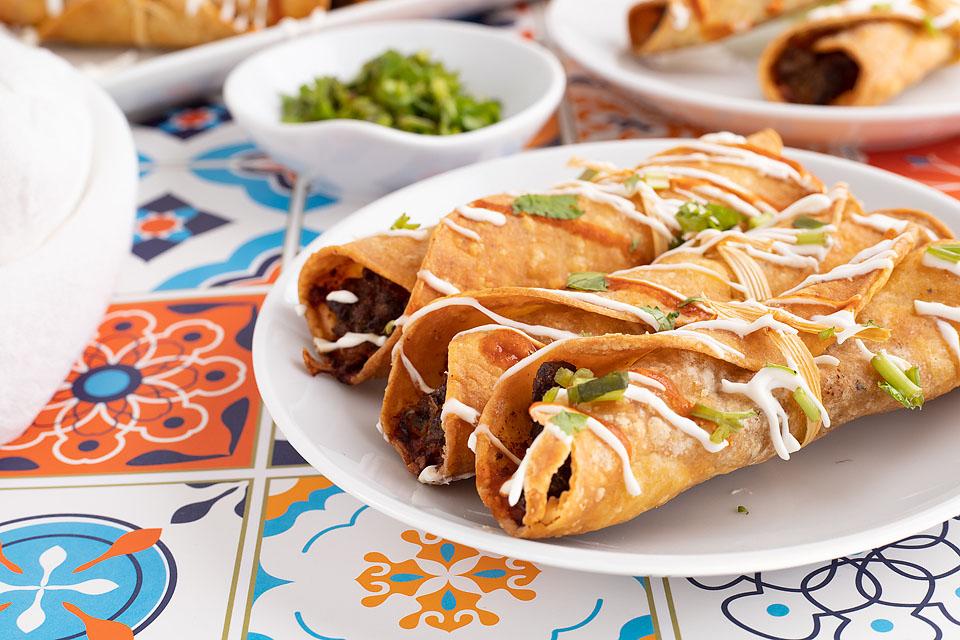 A plate of crispy rolled tacos topped with drizzled white sauce and chopped green herbs, with a small bowl of cilantro in the background on a colorful tiled table.
