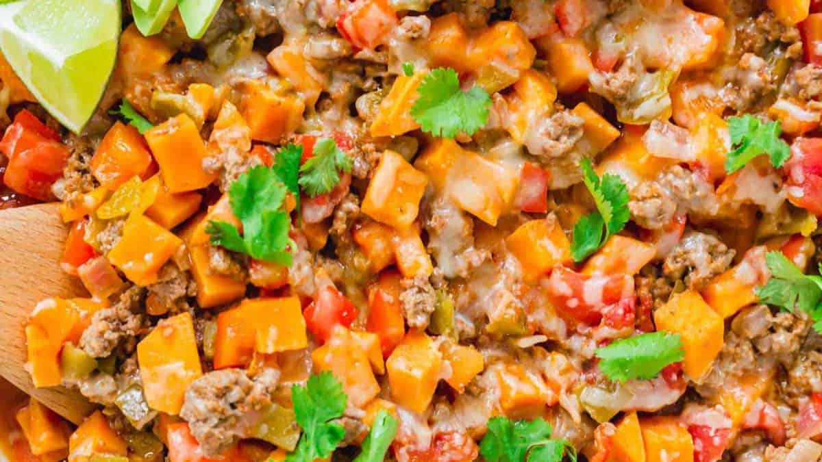 Close-up of a colorful dish with ground meat, diced sweet potatoes, red and green peppers, melted cheese, cilantro, and lime wedges.