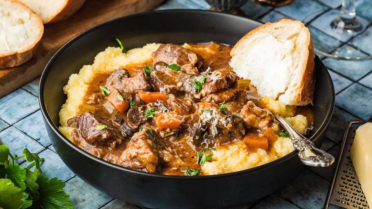 A bowl of beef stew with vegetables served over creamy polenta, garnished with herbs, accompanied by a slice of bread.