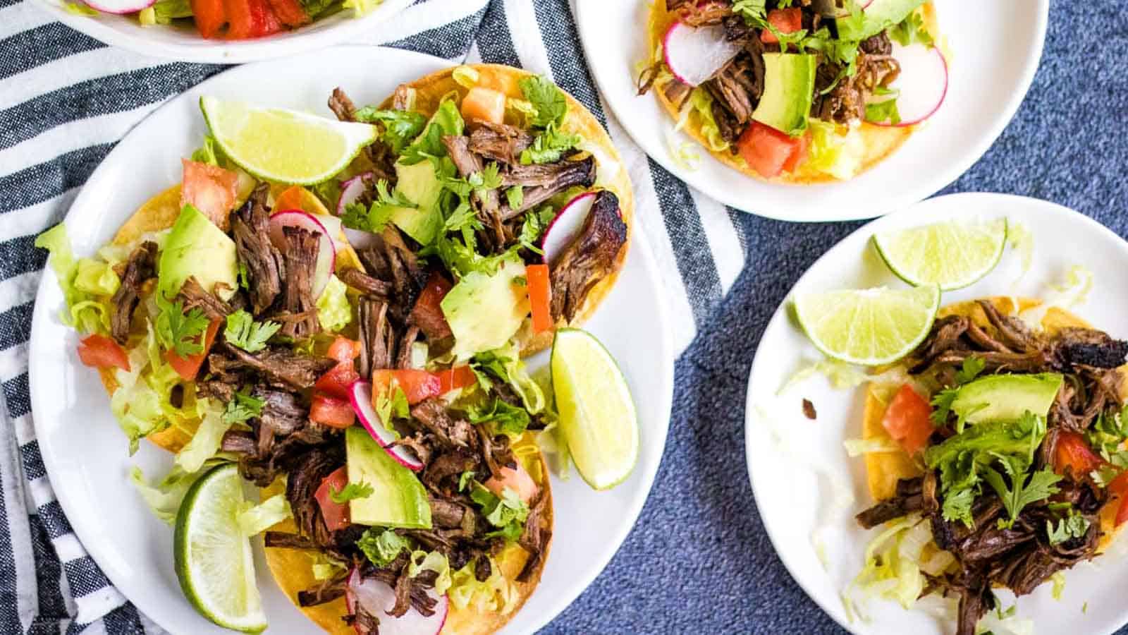 Three plates of tostadas topped with shredded beef, avocado, tomatoes, lettuce, radish slices, cilantro, and lime wedges on a striped cloth.