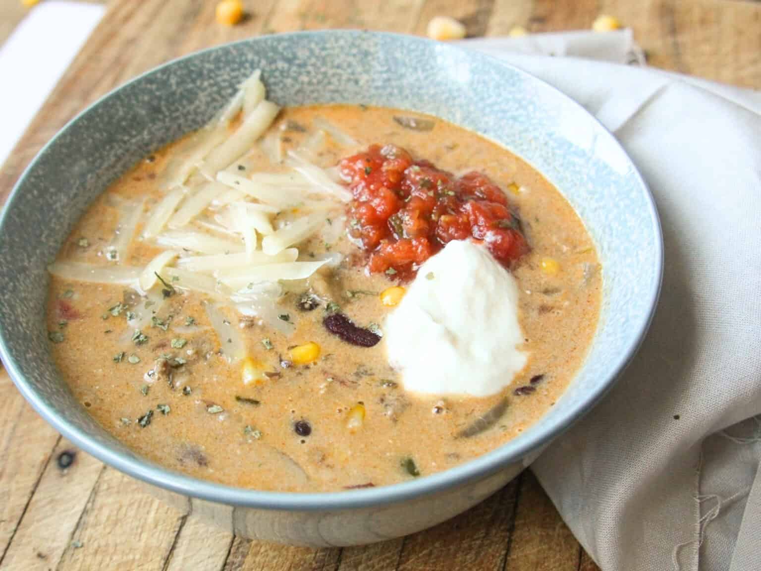 A bowl of creamy soup containing corn, beans, and herbs, topped with shredded cheese, salsa, and a dollop of sour cream, placed on a wooden surface next to a beige napkin.