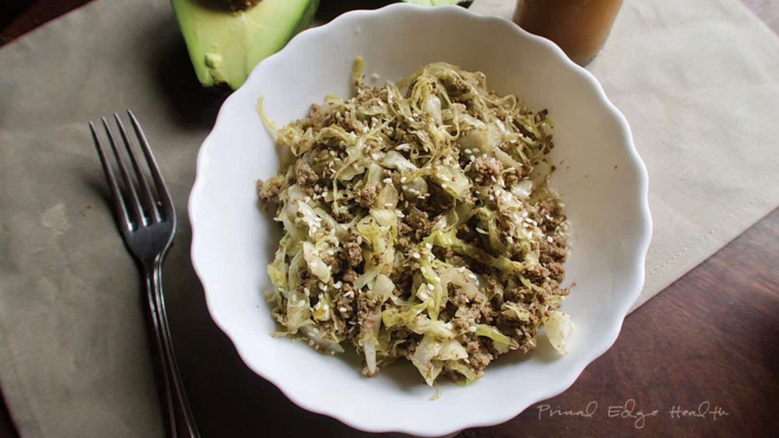 A white bowl filled with shredded cabbage and ground meat, garnished with sesame seeds. A fork and halved avocado are placed nearby on a cloth napkin.
