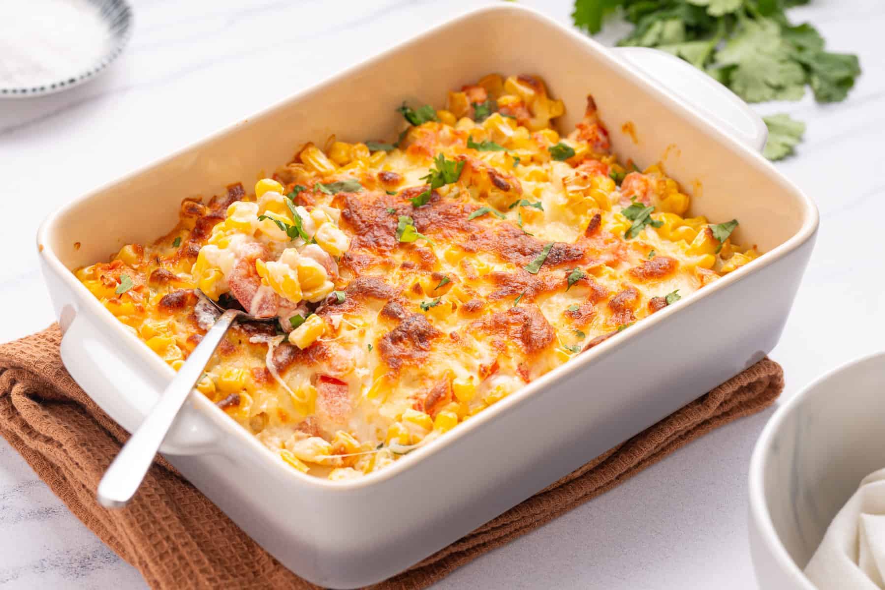 A baked casserole dish filled with corn, cheese, and herbs, with a serving spoon inside and garnished with chopped parsley.