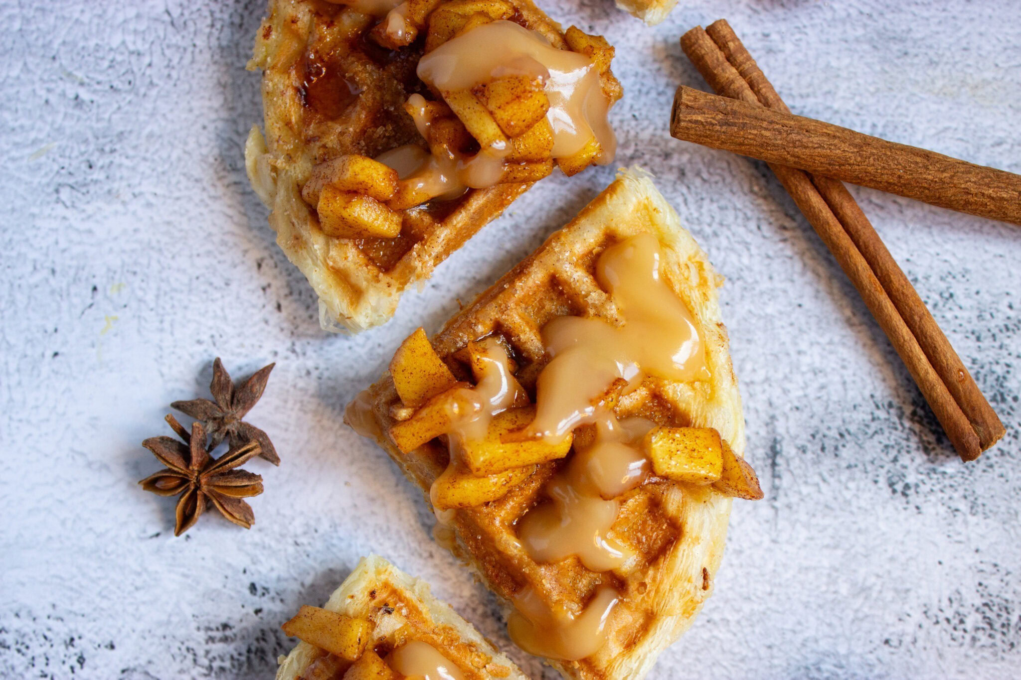 Waffles topped with caramelized apples and sauce, garnished with star anise and cinnamon sticks on a light textured surface.