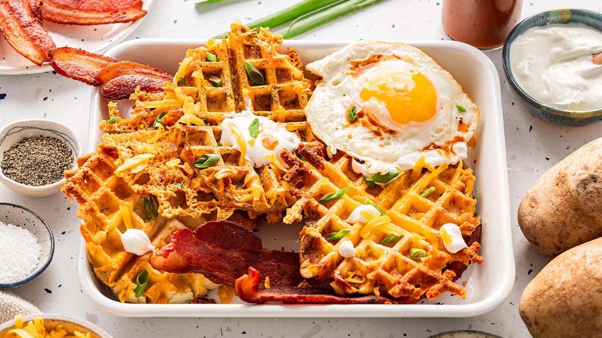 A plate with waffle-shaped hash browns, crispy bacon strips, a sunny-side-up egg, sour cream, and green onions, surrounded by potatoes and condiments.