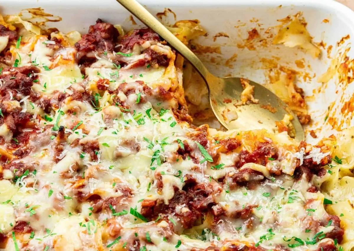 A partially served baked pasta casserole topped with melted cheese, tomato sauce, and herbs in a white dish with a serving spoon.