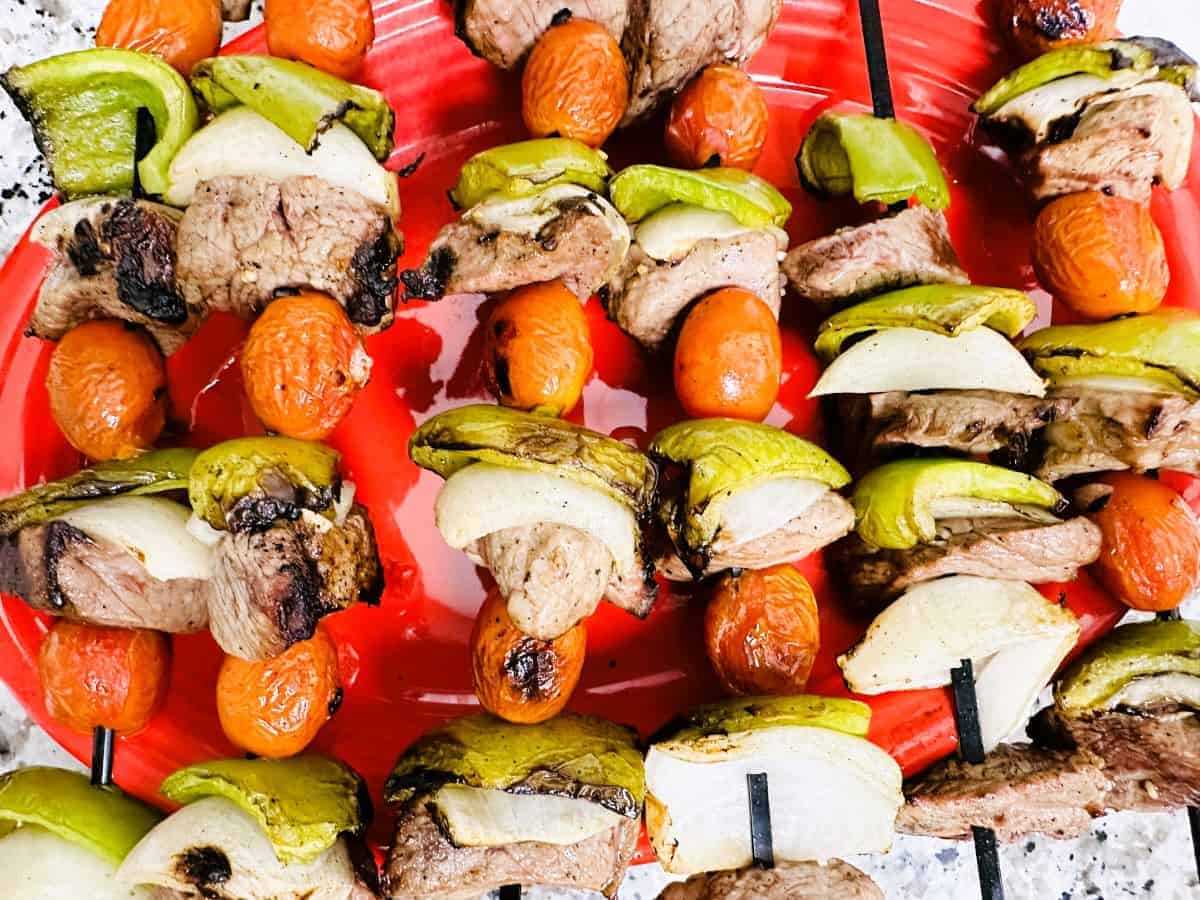 Skewers with grilled beef, green bell peppers, onions, and cherry tomatoes arranged on a red plate.