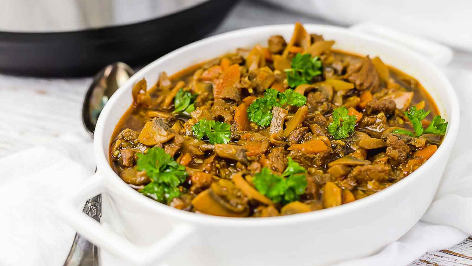 A white dish filled with a stew containing beef, mushrooms, and carrots, garnished with fresh parsley, sits on a white surface next to a spoon.