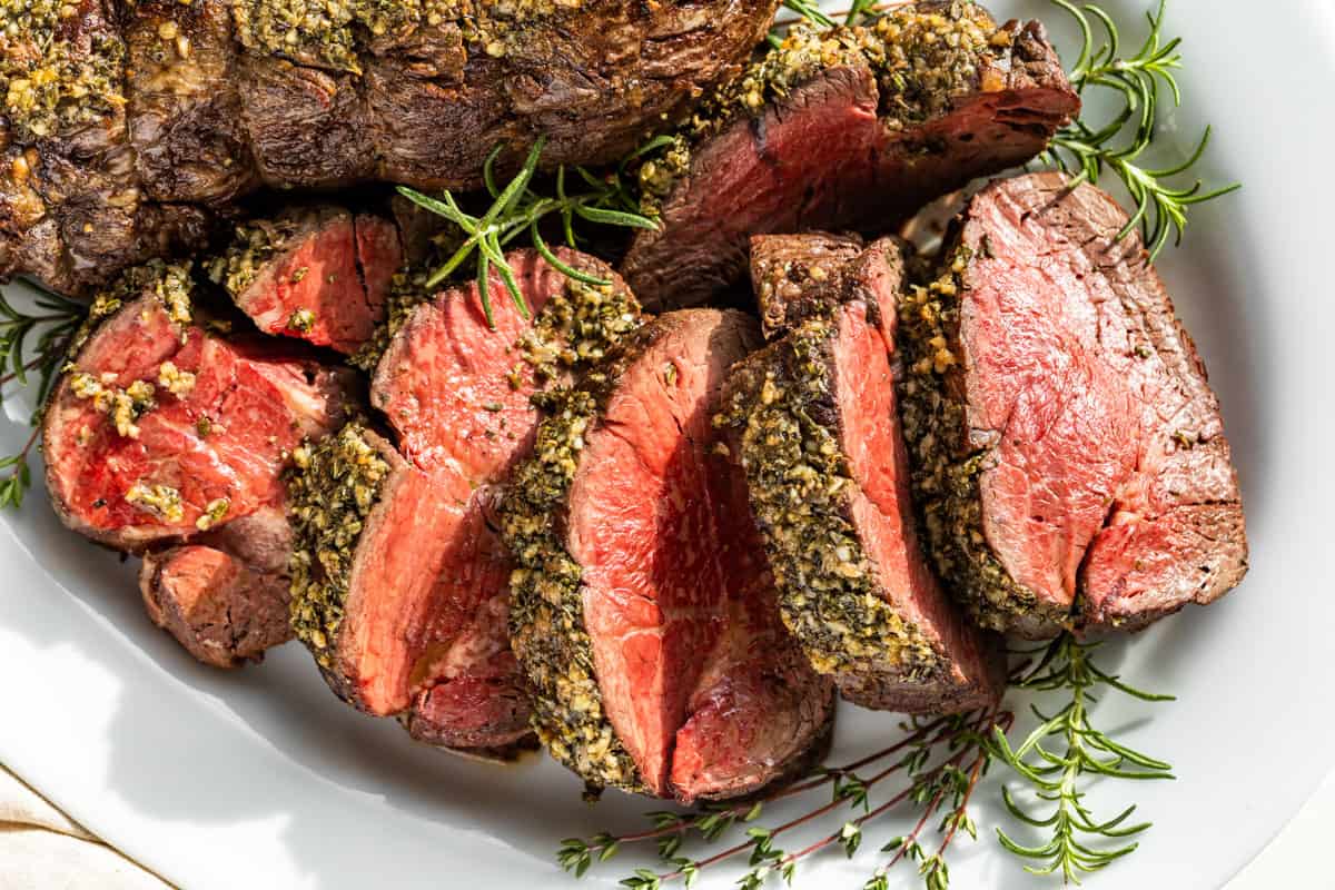 Sliced herb-crusted beef tenderloin served on a white plate with sprigs of fresh rosemary.