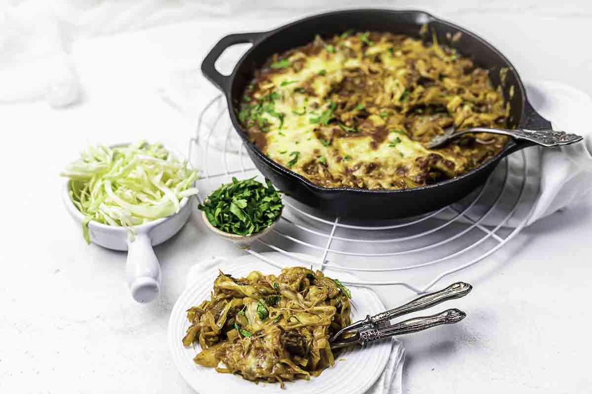 A cast iron skillet filled with baked, cheesy pasta sits on a cooling rack; a serving is plated beside bowls of shredded cabbage and chopped herbs.