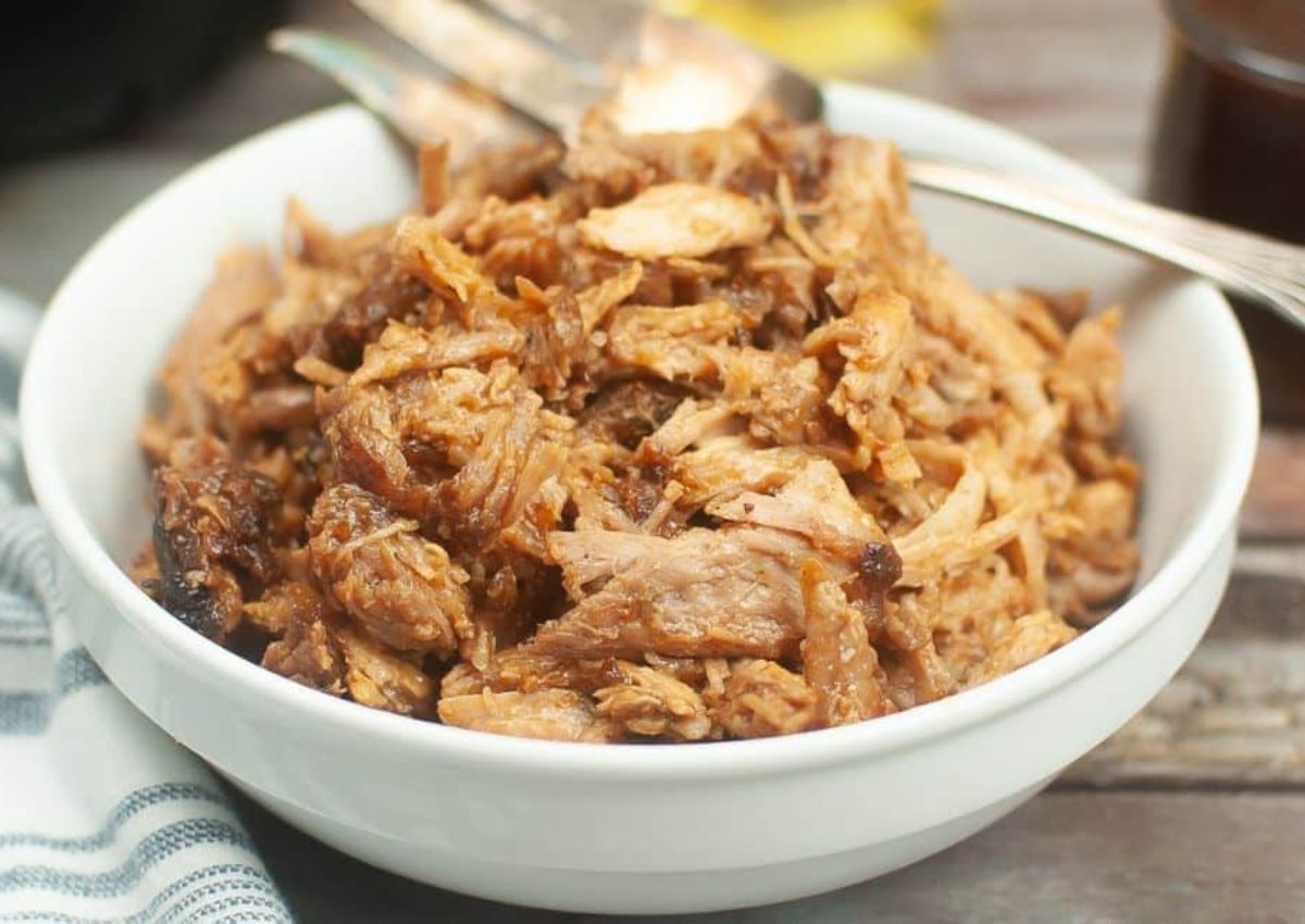 A white bowl filled with shredded, cooked pork sits on a table next to a metal serving fork and tongs.