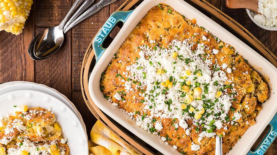 A baked corn casserole in a dish, topped with crumbled cheese, chopped herbs, and corn kernels, with a serving plated nearby. Spoons and fresh corn are also visible.