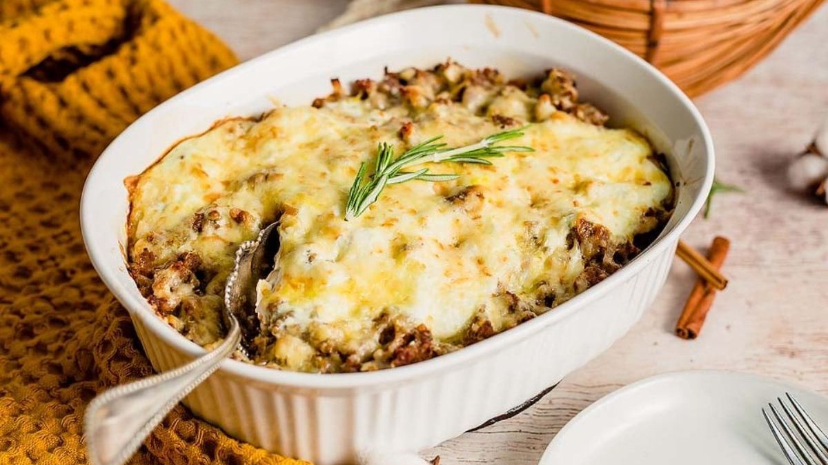A baked casserole with a golden cheese topping in a white dish, garnished with rosemary; a serving spoon is placed inside.
