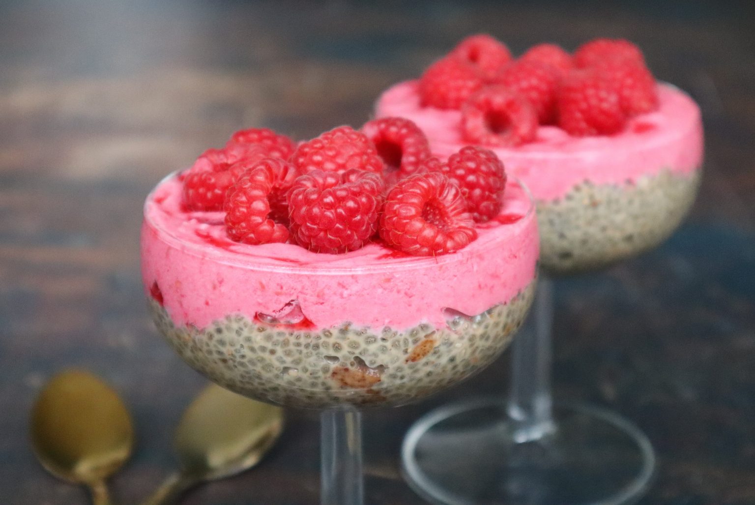 Two glass cups filled with chia pudding, topped with pink raspberry cream and fresh raspberries, with two spoons in the background.