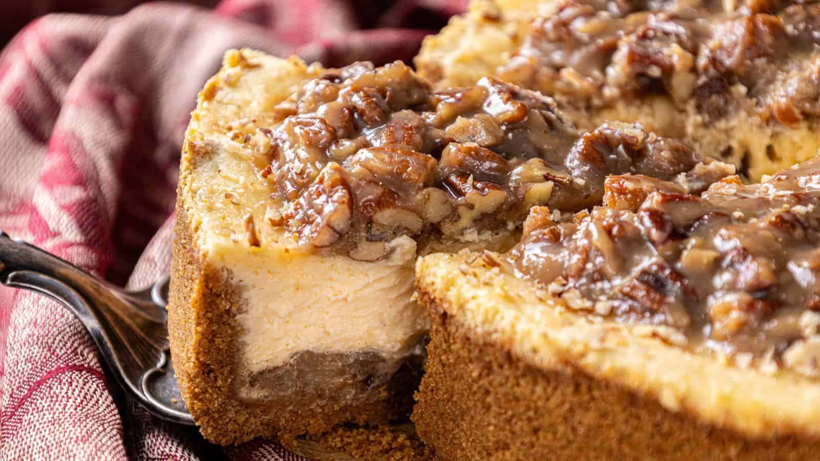 A close-up of a sliced pecan cheesecake with a graham cracker crust, topped with chopped pecans and glaze, on a cloth-covered surface.